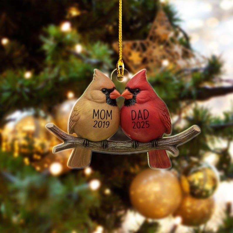 Puede incluir: Adorno navide&ntilde;o con dos cardenales posados en una rama. Un p&aacute;jaro es de color beige con la palabra "MOM" y el a&ntilde;o "2019", el otro es rojo con la palabra "DAD" y el a&ntilde;o "2025". El adorno cuelga de un hilo dorado.