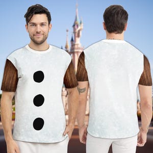 May include: White t-shirt with brown sleeves and three large black circles down the front. The shirt has a light blue speckled pattern and a white collar. The background features a castle.