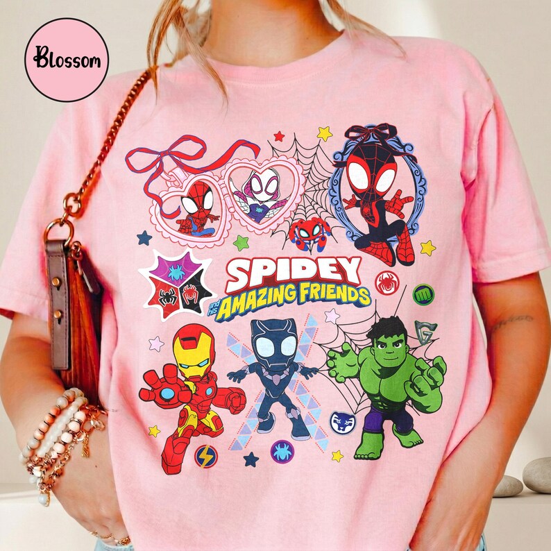 K&ouml;nnte beinhalten: Hellrosa T-Shirt mit dem Schriftzug "Spidey and Amazing Friends" und Cartoon-Illustrationen von Spider-Man, Iron Man, Hulk und Black Panther. Das Shirt hat einen Rundhalsausschnitt und kurze &Auml;rmel.