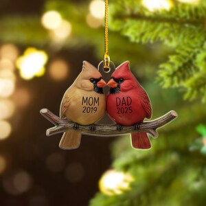 Puede incluir: Adorno navide&ntilde;o decorativo con dos aves cardenales posadas en una rama. Un ave es de color beige con el texto "MOM 2019", y la otra es roja con "DAD 2025". El adorno cuelga de un hilo dorado.