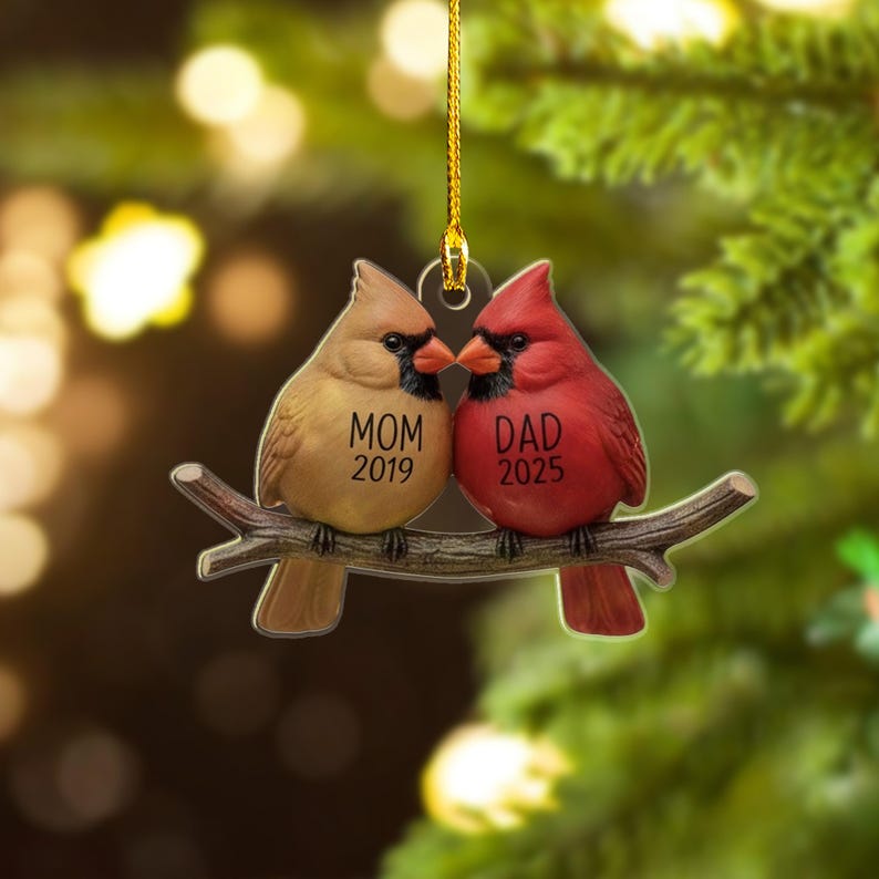 Puede incluir: Adorno navide&ntilde;o decorativo con dos aves cardenales posadas en una rama. Un ave es de color beige con el texto "MOM 2019", y la otra es roja con "DAD 2025". El adorno cuelga de un hilo dorado.