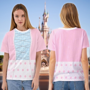 May include: Pink t-shirt with a white crew neck and short sleeves. The front features a light blue corset design with white lace trim and a white panel with pink polka dots. The back is solid pink with a white polka dot panel.
