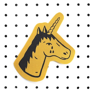 Könnte beinhalten: Ein gelber Einhorn-Aufnäher mit schwarzen Details, einschließlich Mähne, Horn und Tränen. Der Aufnäher befindet sich auf einem weißen Hintergrund mit schwarzen Punkten. Dieser skurrile Aufnäher eignet sich für Kleidung oder Accessoires.