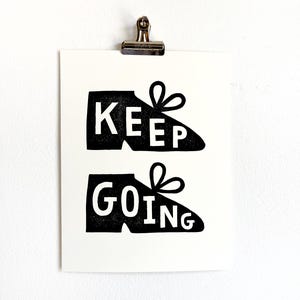 Puede incluir: Impresión en blanco y negro con dos zapatillas deportivas estilizadas y las palabras "KEEP" y "GOING" en letras blancas. La impresión está sujeta con un clip plateado sobre un fondo blanco, ideal para decoración.