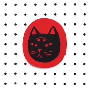 Puede incluir: Un círculo rojo con un gato negro con un tercer ojo en el centro. El gato tiene una expresión seria y mira directamente al espectador.