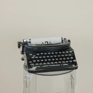 May include: A black vintage typewriter with silver keys and a paper with the text "ΛΟΛ" is displayed on a clear acrylic stand. The typewriter is a classic design, with a glossy finish and a retro aesthetic.