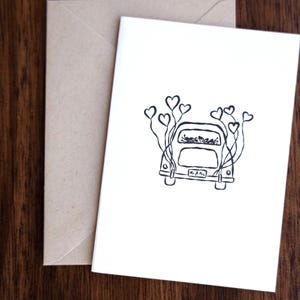May include: A white card with a black line drawing of a car decorated with heart-shaped balloons. The card is next to a tan envelope on a wooden surface. The car has the text "Mr. & Mrs." on the back.