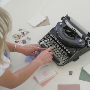 May include: A black vintage typewriter with round keys, surrounded by stationery. The scene includes stamps, envelopes, and notecards. The typewriter is the focal point, suggesting a theme of writing or correspondence.