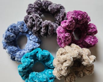 May include: A collection of six plush scrunchies in various colors, including blue, teal, purple, and beige. The scrunchies are made of a soft, velvety material and are arranged in a circular pattern against a white background.