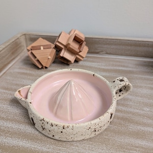 May include: A ceramic citrus juicer with a light pink interior and a speckled white exterior. The juicer has a conical reamer and two small handles. Two wooden puzzles are in the background.
