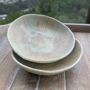 May include: Two ceramic bowls stacked on a wooden surface. The bowls have a light blue and beige color scheme with a speckled interior. The bowls are oval-shaped and have a glossy finish. The background shows a blurred view of trees.