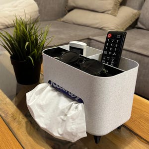 May include: White tissue box holder with a gray speckled finish and black legs. The holder has three compartments for storing items like remotes, sunglasses, and other small items. The holder is sitting on a wooden table.