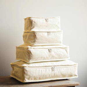 Peut inclure: Une pile de quatre cubes de rangement en toile écrue avec fermetures éclair. Les cubes sont rectangulaires, avec une fermeture éclair sur le dessus. Ils sont empilés, le plus grand en bas et le plus petit en haut.