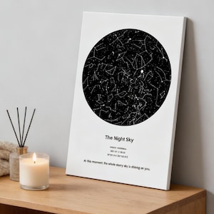 Puede incluir: Impresión en lienzo blanco con un mapa estelar en blanco y negro en un diseño circular. El texto "The Night Sky" está debajo del mapa estelar, junto con texto adicional. Una vela encendida y un difusor están en primer plano.