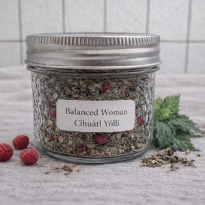 May include: A glass jar with a silver lid, filled with dried herbs and berries. The label reads "Balanced Woman Cihuātl Yōlli". Fresh raspberries and green herbs are scattered around the jar.