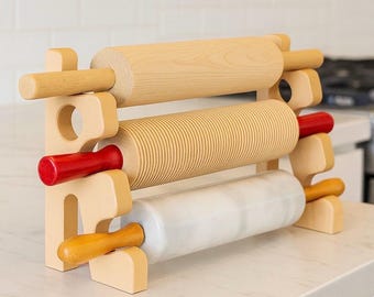 Könnte beinhalten: Ein Holz-Nudelholzständer mit drei Nudelhölzern. Der Ständer ist aus hellem Holz gefertigt und hat eine rechteckige Form. Die Nudelhölzer sind aus Holz und Marmor mit roten und orangefarbenen Griffen.