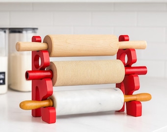 Könnte beinhalten: Ein roter Holz-Nudelholzhalter mit drei Nudelrollen. Die Nudelrollen sind aus Holz, gerilltem Holz und Marmor gefertigt. Der Halter steht auf einer weißen Arbeitsplatte in einer Küche.