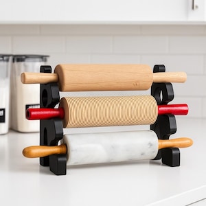May include: A black rolling pin holder with three rolling pins. The top rolling pin is light wood, the middle one is ridged with red handles, and the bottom one is marble with light wood handles. The holder is on a white countertop.