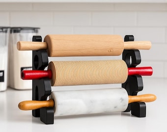 Könnte beinhalten: Ein schwarzer Nudelholzhalter mit drei Nudelrollen. Die obere Nudelrolle ist aus hellem Holz, die mittlere ist geriffelt mit roten Griffen und die untere ist aus Marmor mit hellen Holzgriffen. Der Halter steht auf einer weißen Arbeitsplatte.