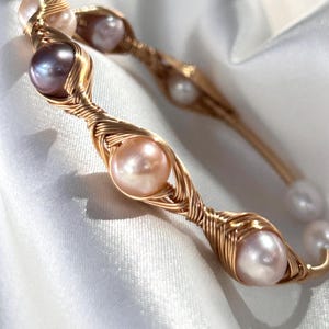 May include: Two gold-toned wire-wrapped bracelets with freshwater pearls. The pearls are in shades of pink, lavender, and white. The bracelets are displayed on a white satin background.