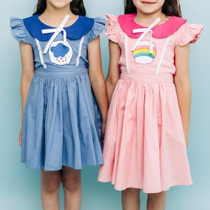 Puede incluir: Dos niñas con vestidos a juego. Uno es azul claro con un gráfico de nube y lluvia, el otro es rosa con un gráfico de arcoíris. Ambos vestidos tienen un cuello en contraste y un lazo blanco.