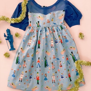 Puede incluir: Un vestido azul claro con un patrón repetitivo de cascanueces, bailarines y dulces navideños. La prenda tiene mangas abullonadas cortas y transparentes de color azul oscuro y un canesú a juego. Una pequeña figura de cascanueces azul se encuentra a la izquierda.