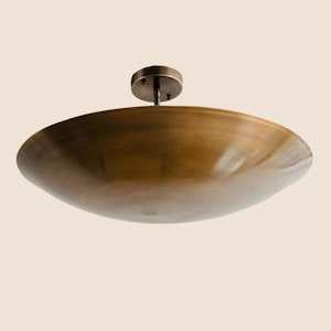 Puede incluir: Una lámpara de techo en forma de cuenco poco profundo de color bronce. La lámpara tiene una placa de montaje circular y una pantalla curva y ancha. El diseño general es minimalista y moderno.