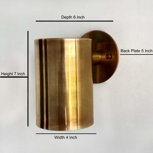 Könnte beinhalten: Eine goldfarbene Wandleuchte mit einem zylindrischen Schirm. Die Leuchte ist 17,8 cm hoch, 10,2 cm breit und 15,2 cm tief. Die Rückplatte hat einen Durchmesser von 12,7 cm.