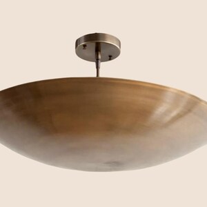 May include: A bronze-colored, shallow, circular ceiling light fixture. The fixture has a wide, bowl-shaped shade and a small, round mounting plate. The light is suspended from a short rod.