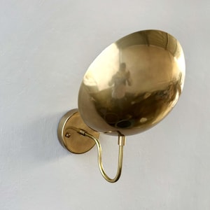 Op de afbeelding: Een messing wandlamp met een ronde, gebogen kap. De kap is naar beneden gericht en heeft een glanzend, reflecterend oppervlak.
