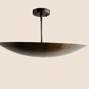 May include: A bronze-colored, modern pendant light fixture. The light has a wide, shallow, bowl-shaped shade suspended from a central rod and a circular ceiling mount. The fixture is designed for overhead illumination.