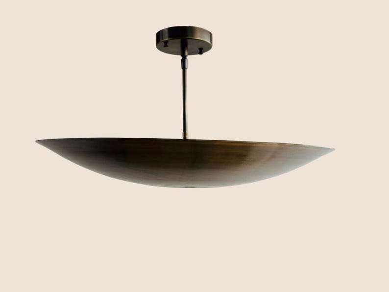 May include: A bronze-colored, modern pendant light fixture. The light has a wide, shallow, bowl-shaped shade suspended from a central rod and a circular ceiling mount. The fixture is designed for overhead illumination.