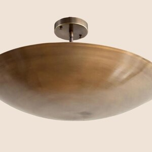 May include: A bronze-colored, bowl-shaped ceiling light fixture. The fixture has a circular base and a stem that connects to the bowl. The bowl has a smooth, curved surface. The light fixture is designed to be mounted on a ceiling.