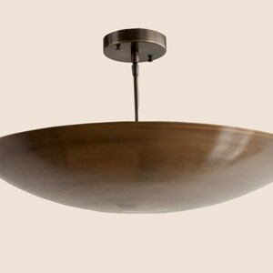 May include: A bronze-colored, bowl-shaped ceiling light fixture. The fixture has a circular base and a slender rod suspending the bowl. The bowl has a wide, shallow design. The light fixture is designed to be mounted on a ceiling.