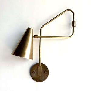 Può includere: Un'applique da parete in ottone con un paralume a forma di cono e un braccio geometrico. L'applique ha un design minimalista e aggiungerebbe un tocco moderno a qualsiasi stanza.