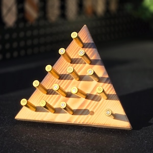 Può includere: Espositore triangolare in legno con bossoli in ottone disposti in file. Il legno è marrone chiaro con venature visibili e i bossoli in ottone hanno una tonalità dorata. L'espositore è progettato per contenere oggetti.