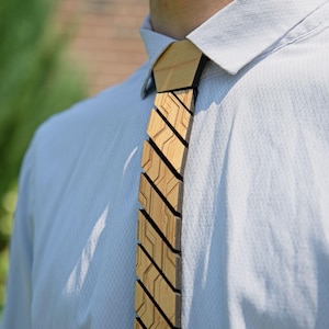 May include: A wooden tie with a geometric design. The tie is made of light-coloured wood and features a series of angled, rectangular segments. The tie is worn over a light blue dress shirt.