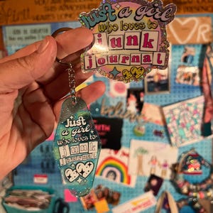 May include: A keychain and a sticker with the text "Just a girl who loves to junk journal." The keychain is a teal motel-style tag with glitter and hearts. The sticker has a pink, purple, and teal color scheme with a holographic border.
