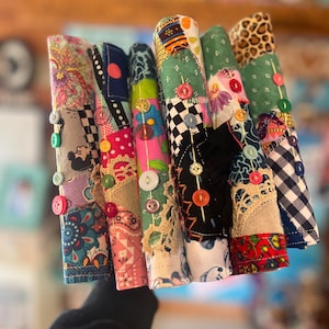 May include: A collection of colorful, fabric-covered items, each adorned with decorative buttons. The fabrics feature various patterns, including floral, paisley, and checkered designs. The items are rolled up, showcasing the diverse textures and colors.