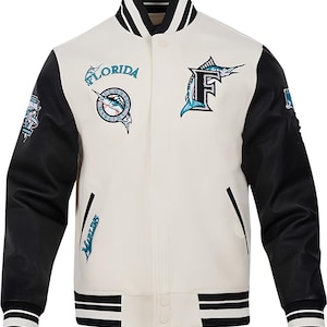 May include: Cream-colored varsity jacket with black sleeves and striped cuffs. Features the Florida Marlins logo and lettering in teal and white. Includes a button-up front and two front pockets.