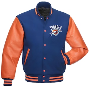 Może przedstawiać: Niebiesko-pomarańczowa kurtka typu varsity z napisem "THUNDER OKC" na piersi. Kurtka ma pomarańczowe skórzane rękawy, niebieski korpus oraz pomarańczowo-niebieskie paski na mankietach i u dołu. Zapinana na guziki, z dwiema kieszeniami.