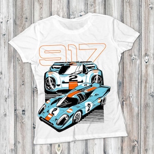 Pode incluir: Camiseta branca com um gráfico de dois carros de corrida vintage em azul, laranja e preto. O número "2" é exibido nos carros. A palavra "917" está impressa em laranja acima dos carros.
