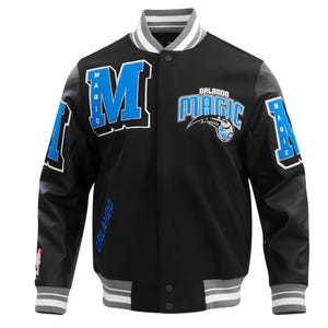 Op de afbeelding: Zwarte en grijze letterman-jas met het Orlando Magic-logo. De jas toont de naam en het logo van het team in blauw en wit, met de letter "M" op de voorkant. De mouwen zijn grijs met zwart leerachtig materiaal.