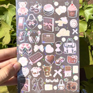 May include: A sheet of holographic stickers featuring various designs, including a pink teddy bear, cupcakes, bows, handbags, and chocolate bars. The stickers have a pastel color palette with glitter accents, creating a cute and whimsical aesthetic.