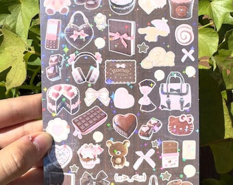 Peut inclure: Une feuille d'autocollants holographiques avec divers motifs, dont un ours en peluche rose, des cupcakes, des nœuds, des sacs à main et des barres chocolatées. Les autocollants ont une palette de couleurs pastel avec des accents de paillettes.