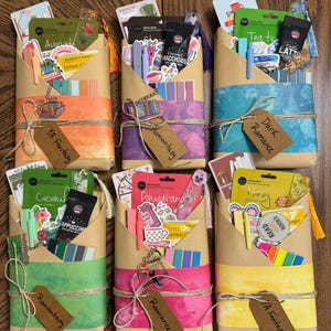 May include: Nine wrapped packages with different colored paper and twine. Each package has a small tag with a different genre of book written on it. The genres include YA Fantasy, Romantasy, Dark Romance, Romance, and Contemporary.