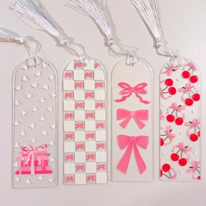 May include: Four clear acrylic bookmarks with white tassels. Each bookmark features a different design: hearts, pink bows on a white grid, pink bows, and pink cherries with bows. A gift box design is also visible.