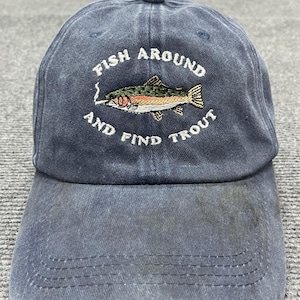 Op de afbeelding: Een vervaagde marineblauwe baseballpet met een borduursel van een forel die een sigaret in de bek houdt. De tekst "FISH AROUND AND FIND TROUT" is in witte draad in een cirkelvormig patroon rond de centrale visillustratie geborduurd.