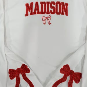Pode incluir: Camisola branca com o nome "MADISON" em letras vermelhas brilhantes no peito. Um laço vermelho é bordado abaixo do nome, com dois laços vermelhos brilhantes adicionais nas mangas. Uma peça de roupa casual e festiva.