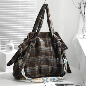 May include: A brown plaid tote bag with blue accents and a patterned shoulder strap. The bag has two handles and a decorative knot detail. The bag is sitting on a white surface.